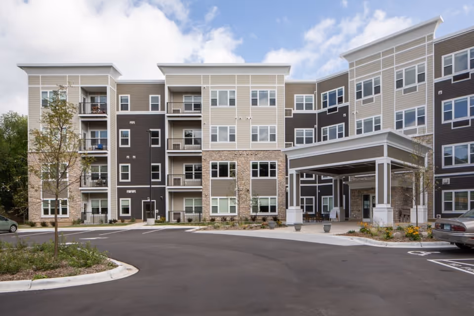 Front exterior of a modern multi-story senior living building with a covered entrance, balconies, and parking area.