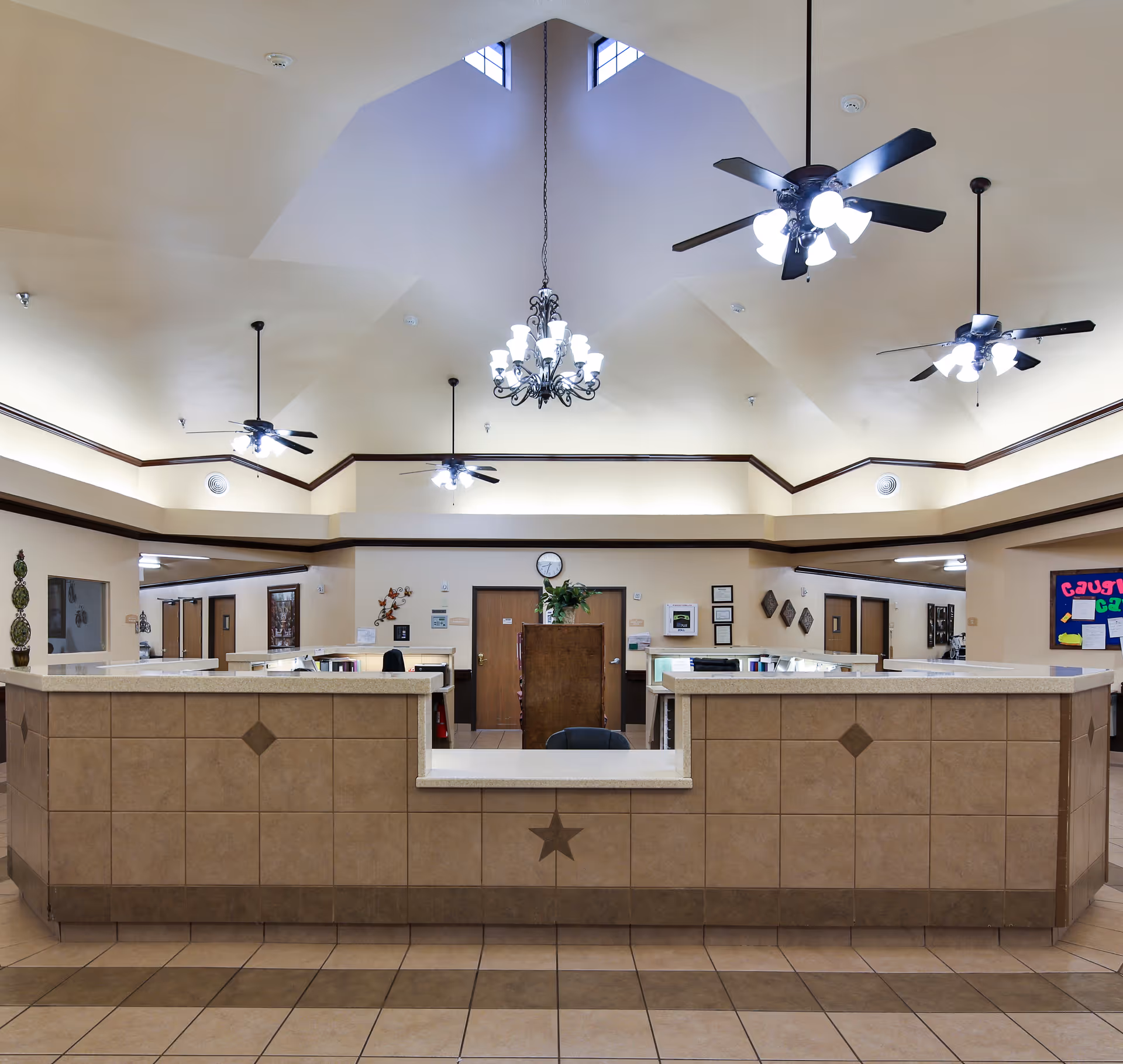 Reception area of a nursing and rehabilitation center with a large tiled front desk featuring a star design, ceiling fans with lights, a chandelier, and beige walls with dark trim. There are doors and bulletin boards visible in the background.