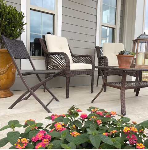 Outdoor patio area with two cushioned wicker chairs, a wicker table with a potted succulent, a folding chair, and a large planter with greenery. Colorful flowers are in the foreground and windows with white trim are visible on the building behind.