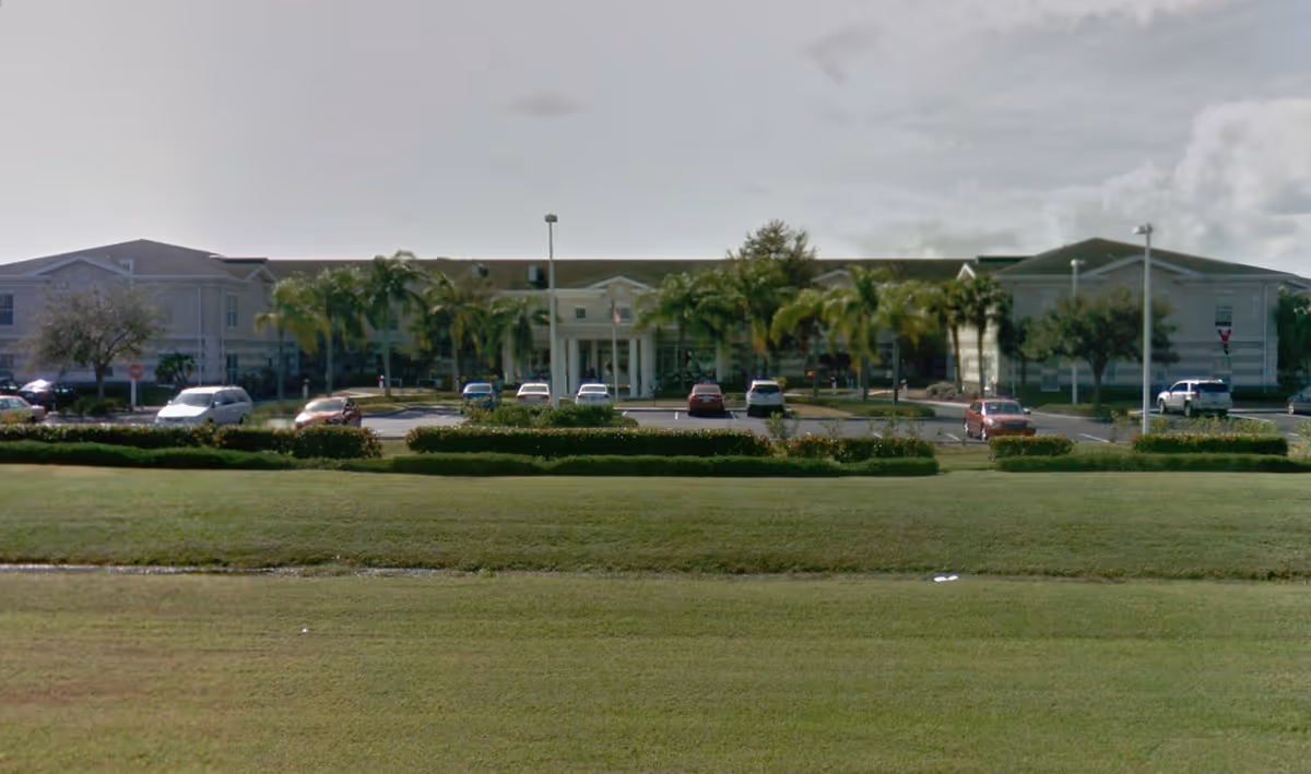 Front exterior view of Sodalis Vero Beach facility with a large lawn in the foreground, a parking lot with several cars, palm trees, and a two-story building under a cloudy sky.