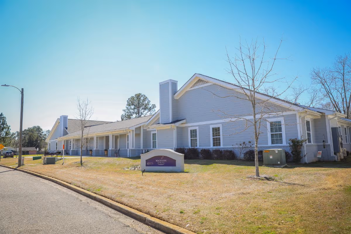 Exterior view of a single-story senior living facility building with light gray siding and white trim. The building has a covered porch with several chairs and a sign in front that reads 'Madison Place'. The sky is clear and blue, and there are a few leafless trees and some shrubs around the building.