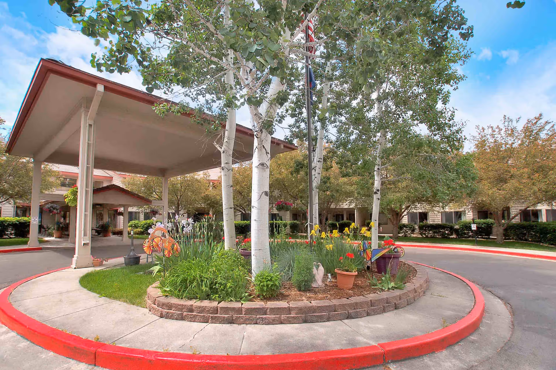 Covered drop-off driveway and circular landscaped island with birch trees and flowers in front of a senior living building.