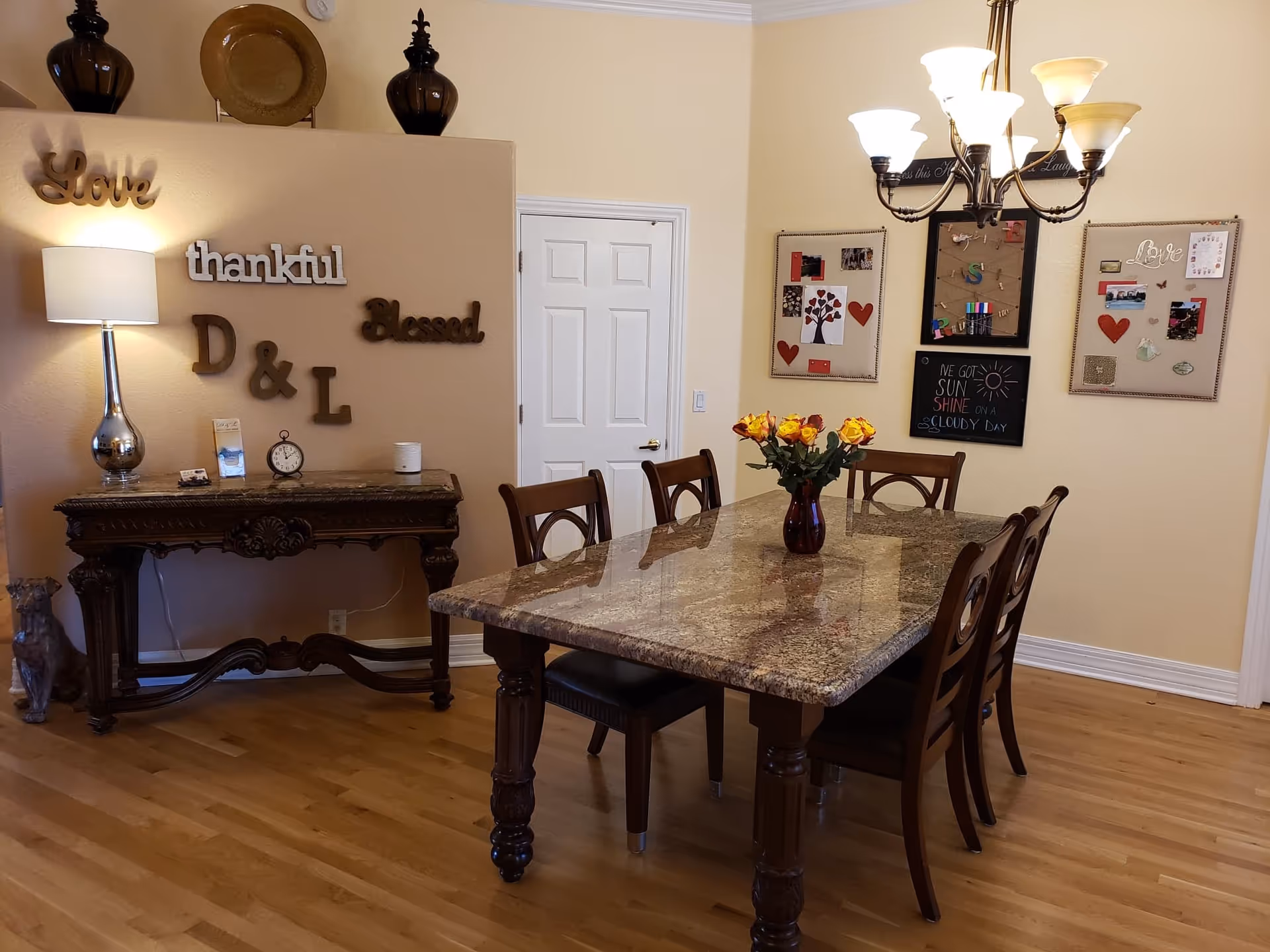 A dining room with a rectangular marble table surrounded by six wooden chairs. A vase with yellow and orange roses is placed in the center of the table. On the left side, there is a wooden console table with a lamp, clock, and decorative items. The wall behind the console table has decorative words including 'Love', 'thankful', 'Blessed', and 'D & L'. On the right wall, there are three bulletin boards with photos, notes, and decorations. A chandelier with six lights hangs above the dining table. The floor is wooden, and the walls are painted beige.