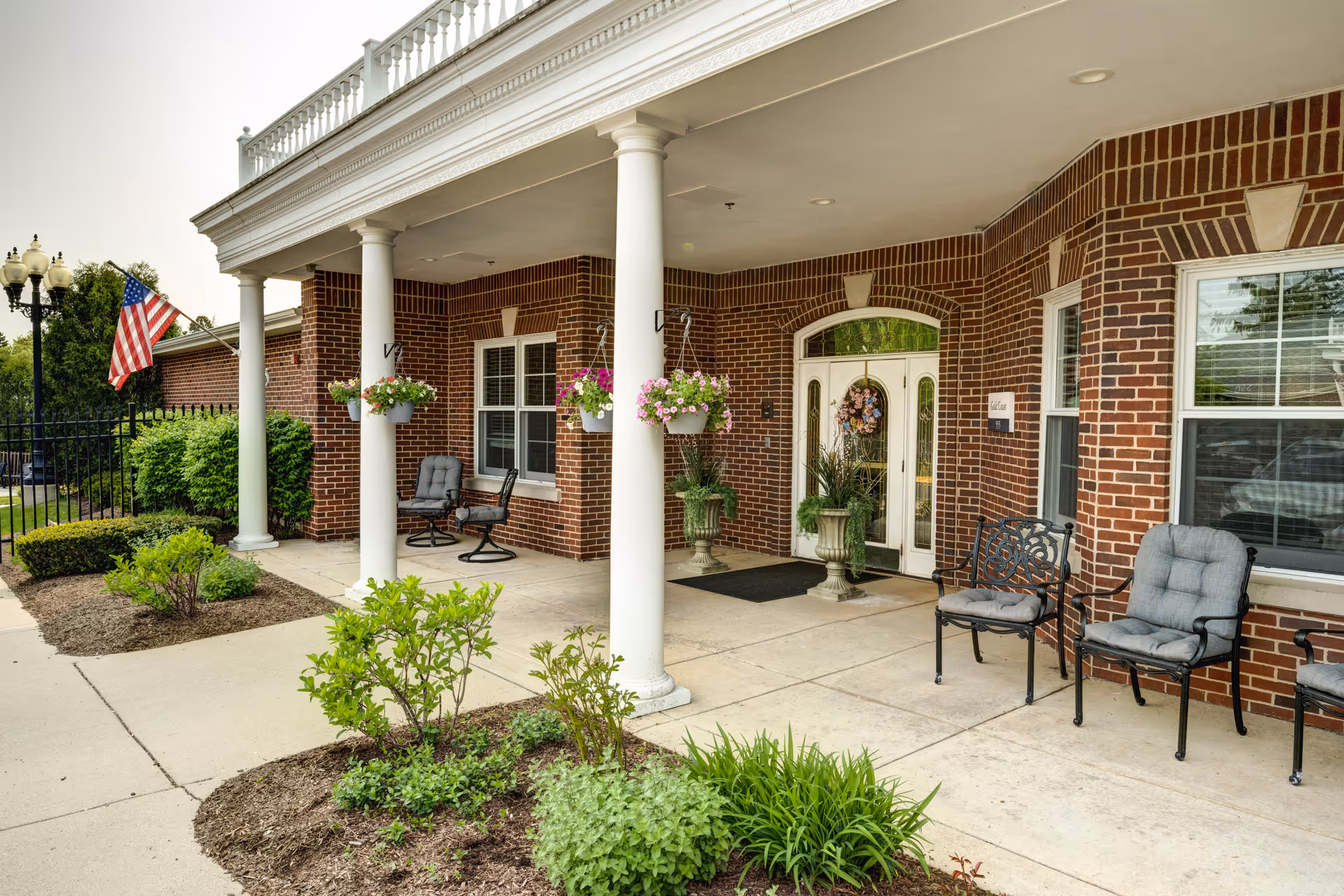 Entrance area of a senior living facility with a brick exterior, white columns, hanging flower pots, cushioned chairs, and an American flag. There are plants and shrubs along the walkway leading to double glass doors decorated with a wreath.