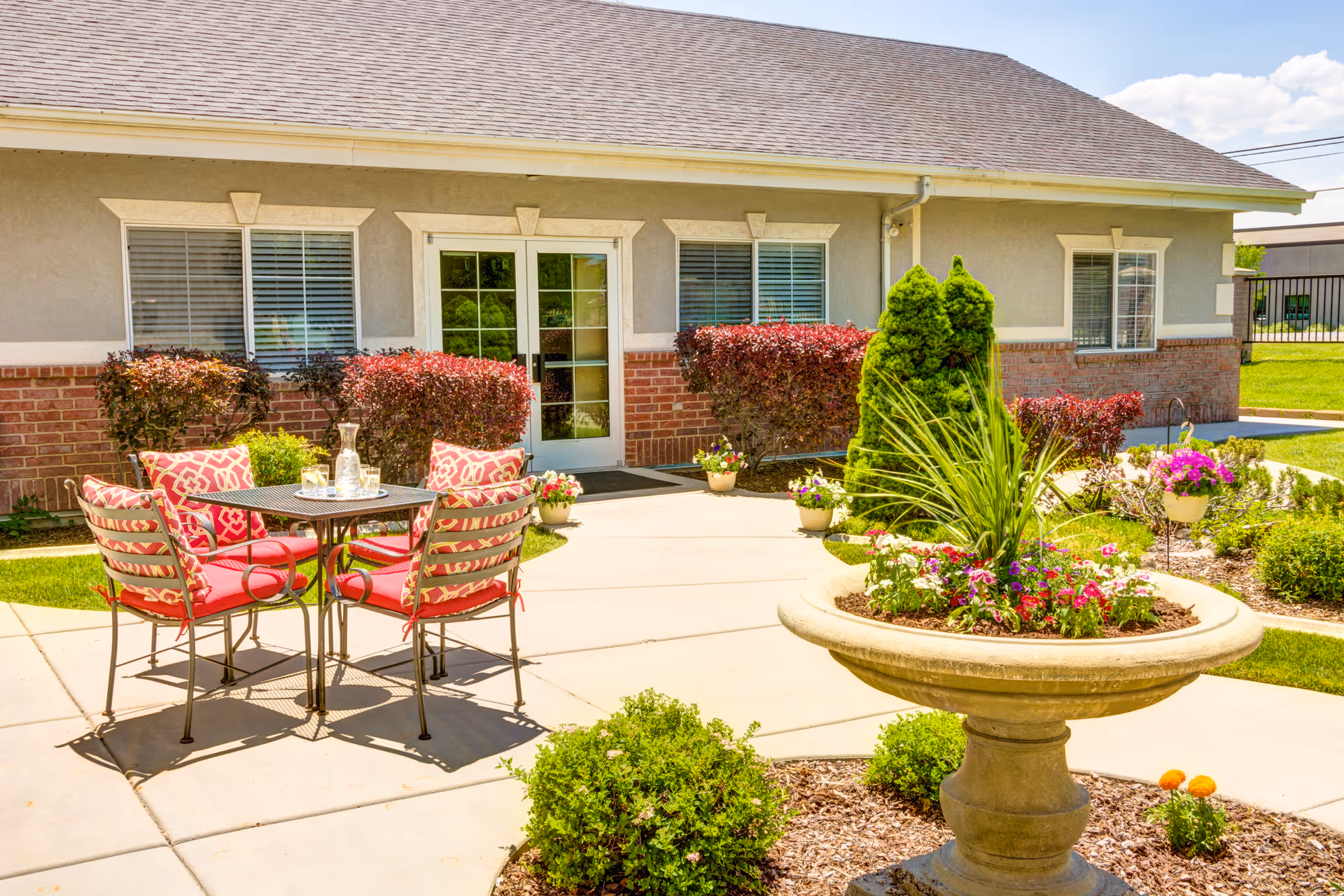 Outdoor patio area with a round metal table and four red cushioned chairs with patterned pillows. The patio is surrounded by well-maintained landscaping including bushes, flowers, and a large decorative planter with greenery. The building in the background has beige walls, brick accents, and multiple windows with white frames.