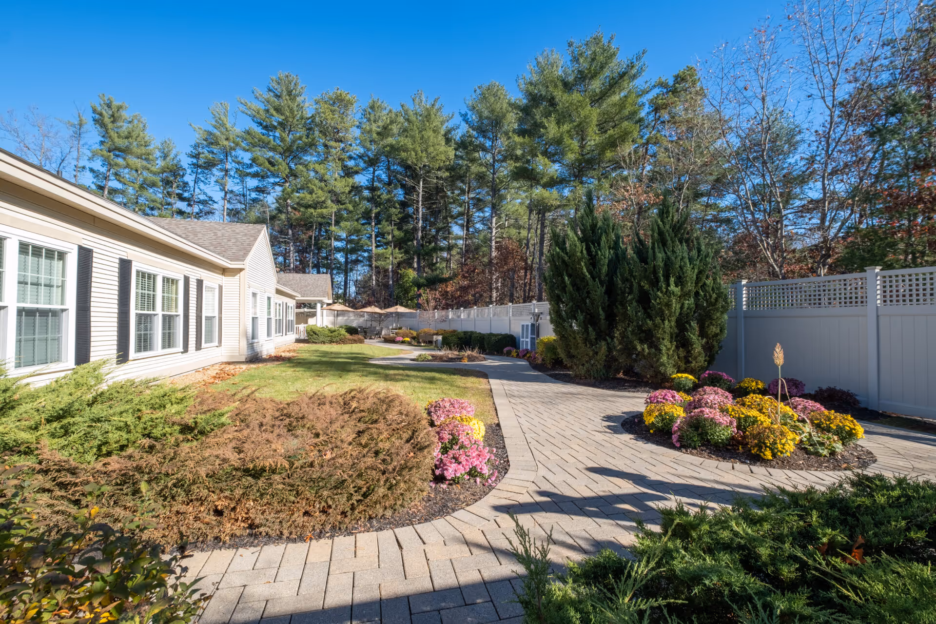 Outdoor garden area at Benchmark Senior Living at Nashua Crossings featuring a paved walkway, landscaped flower beds with colorful flowers, bushes, and trees. The side of a light-colored building with multiple windows is visible on the left, and a white fence runs along the right side of the walkway. Tall pine trees and a clear blue sky are in the background.