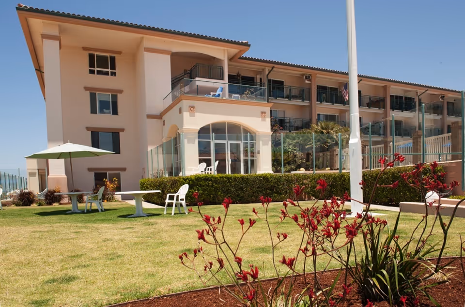Three-story beige senior living building with balconies overlooking a lawn, patio tables and chairs, and red flowers in the foreground under a clear sky.