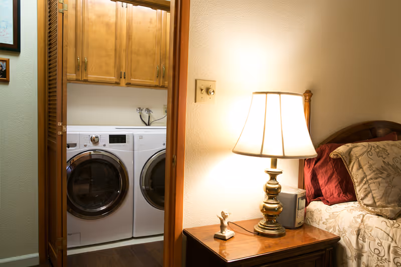A bedroom nightstand with a lit lamp and bed beside an open door revealing a washer and dryer with overhead cabinets.