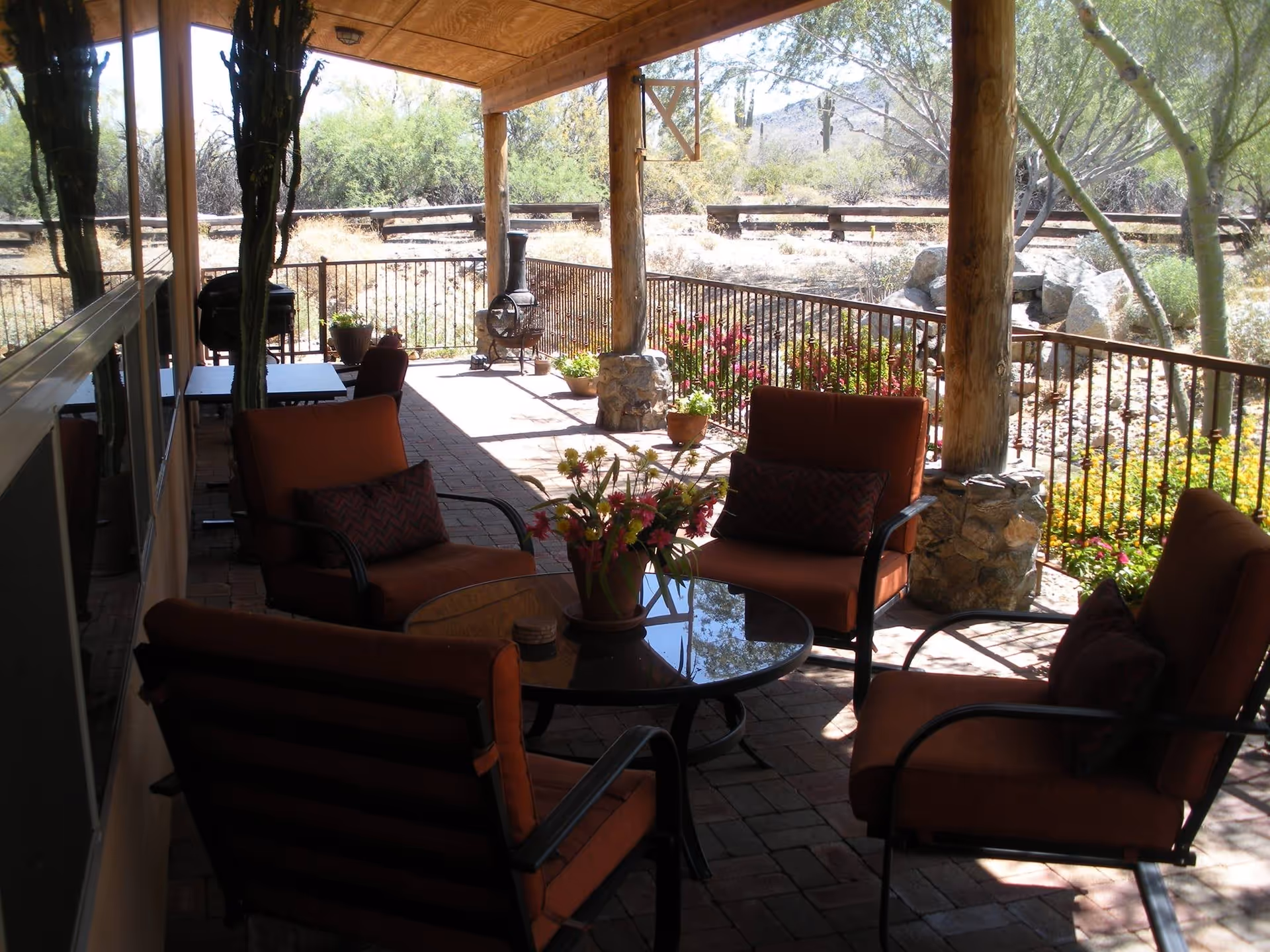 Covered outdoor patio area with four cushioned chairs around a glass-top table with a flower pot centerpiece. The patio has a brick floor, wooden posts, and a metal railing overlooking a garden with flowers and desert plants. There is a barbecue grill and a chiminea in the background.