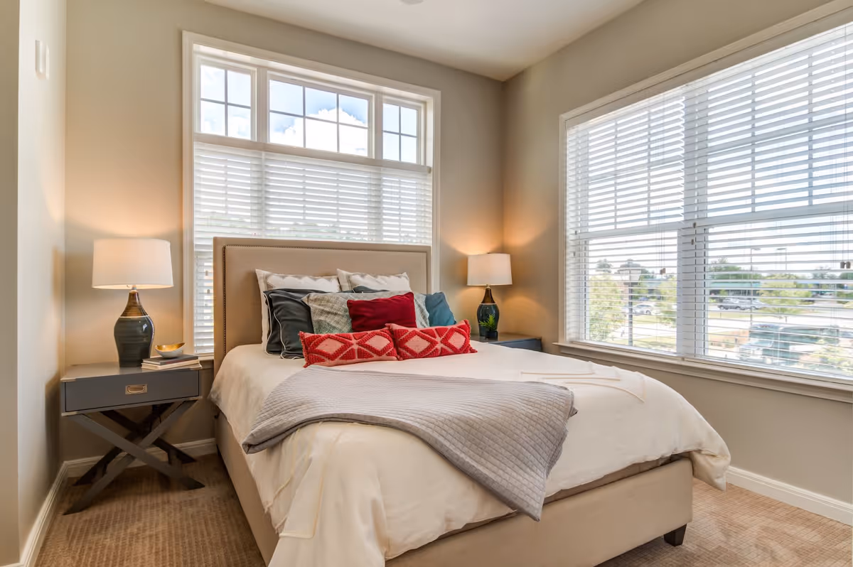 A bright bedroom with a beige upholstered bed adorned with multiple pillows in shades of red, gray, and white. There are two bedside tables, each with a lamp, and large windows with white blinds letting in natural light. The room has beige walls and carpeted flooring.