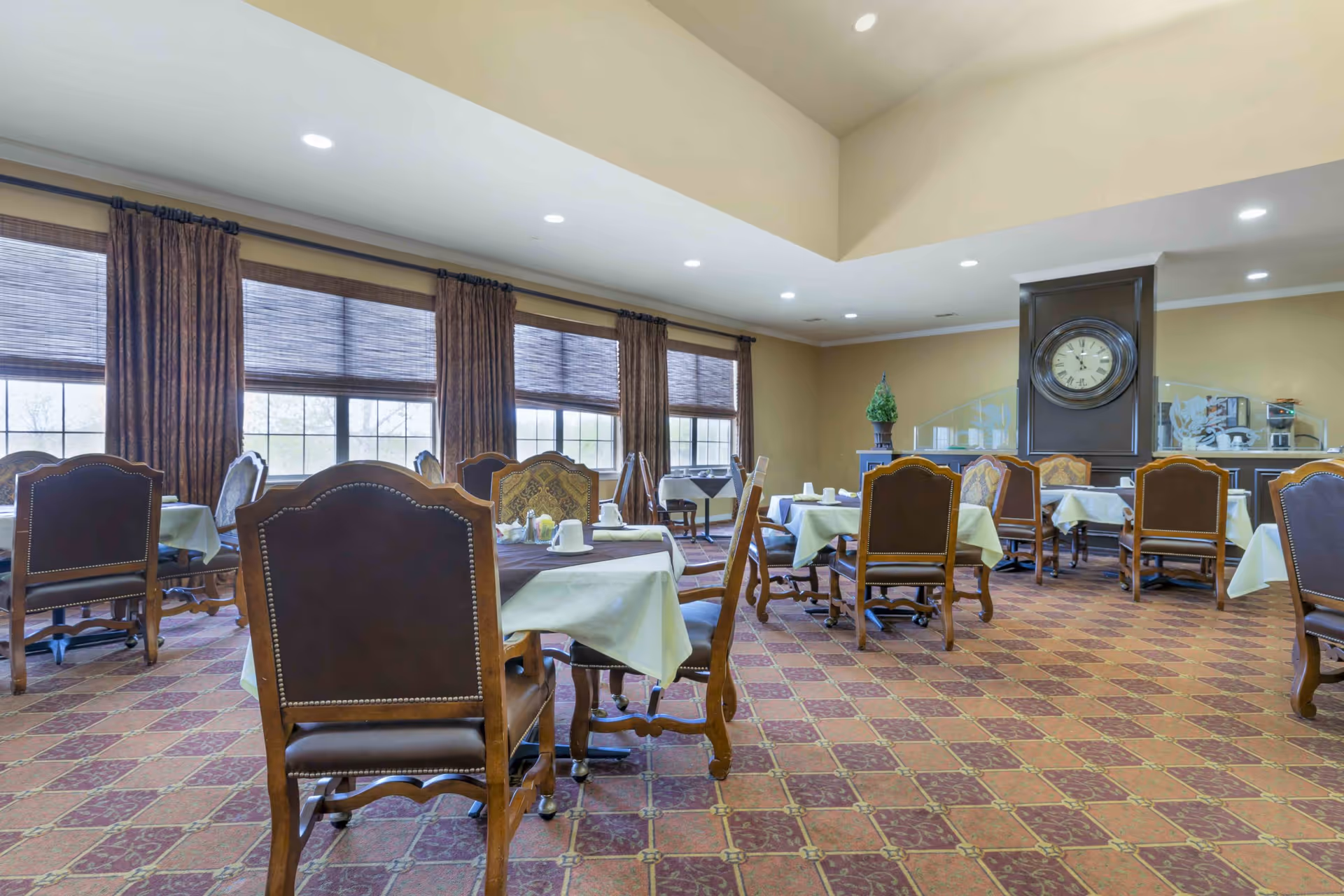 Spacious dining room with multiple tables set with linens and upholstered chairs, large windows with curtains, and a clock on the far wall.