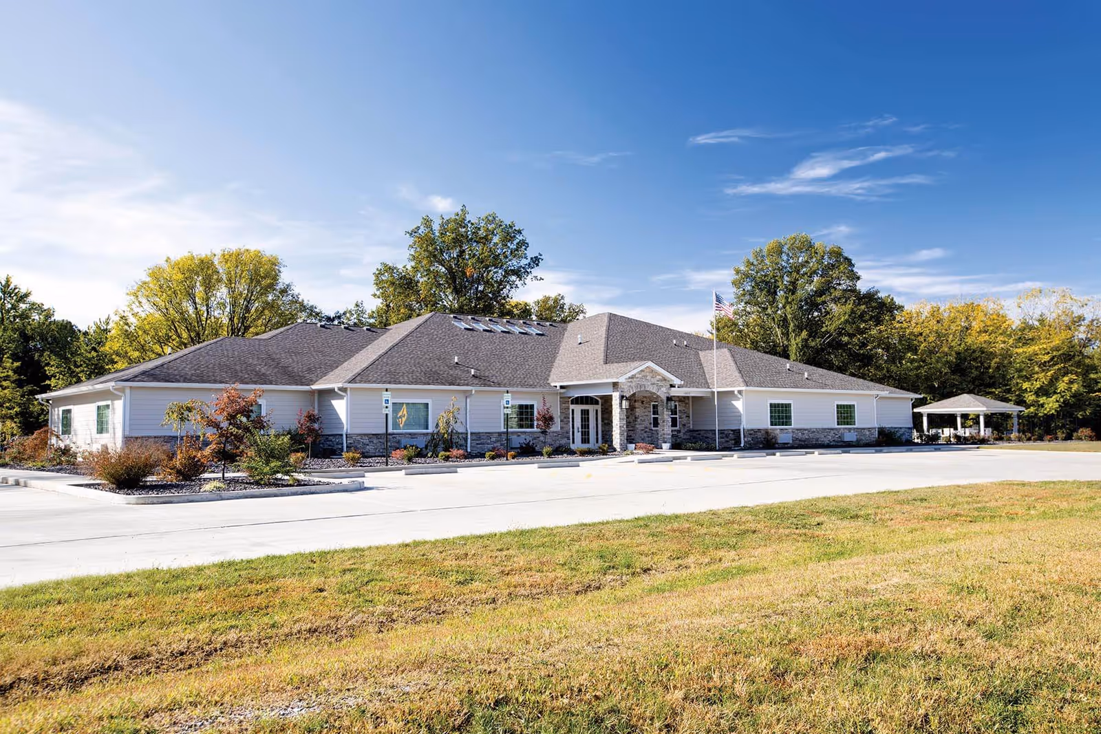 Single-story senior living facility building with a landscaped driveway, American flag, and trees under a clear blue sky.