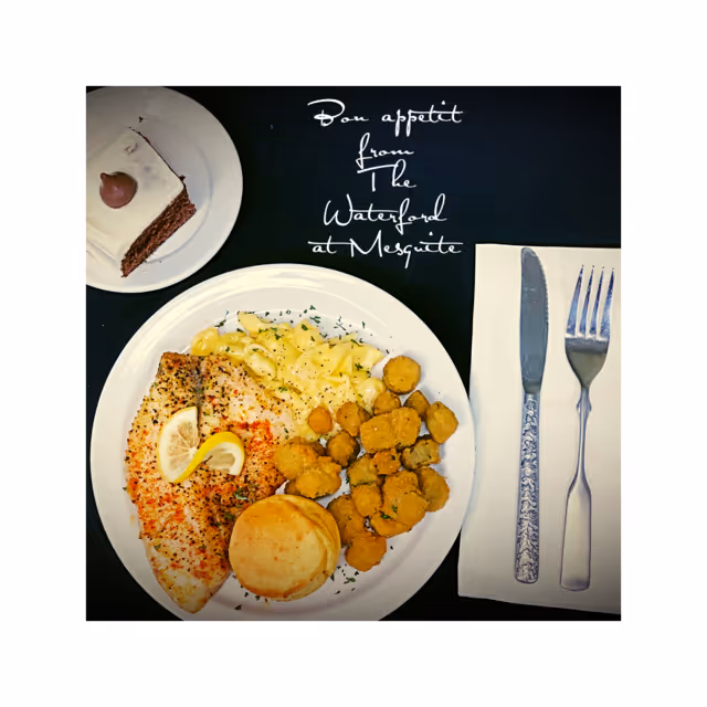 A meal on a white plate consisting of a seasoned fish fillet garnished with lemon slices, macaroni and cheese, fried okra, and a biscuit. Next to the plate is a knife and fork on a white napkin. A slice of chocolate cake with white frosting and a chocolate decoration is on a separate small white plate. The text 'Bon appétit from The Waterford at Mesquite' is written above the meal on a dark background.