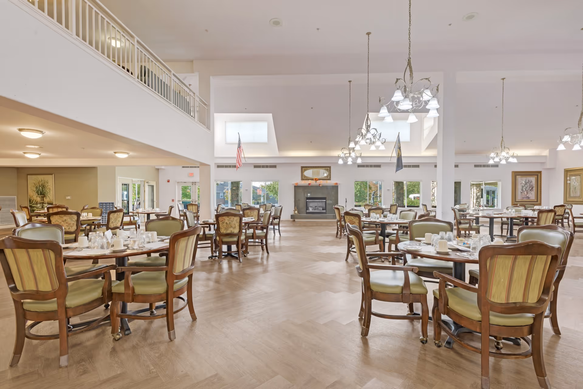 A spacious dining room with multiple round wooden tables and cushioned chairs arranged neatly. The room features large windows allowing natural light, a fireplace in the center, and decorative chandeliers hanging from the ceiling. The floor has a light wood pattern, and there are framed artworks on the walls.