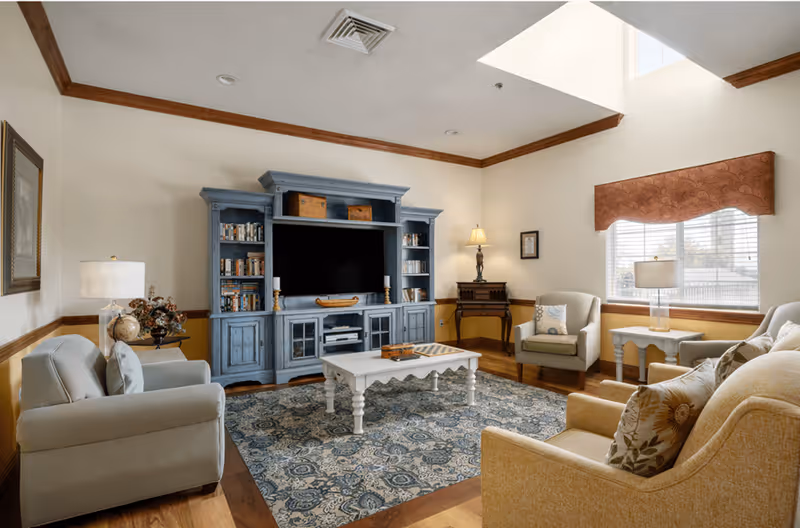 A cozy living room with a large blue entertainment center holding a flat-screen TV and books. The room features a patterned blue and beige rug, a white coffee table, two beige armchairs, a beige sofa with floral pillows, and a side table with a lamp. There is a window with blinds and a valance, and a skylight above allowing natural light to fill the space.