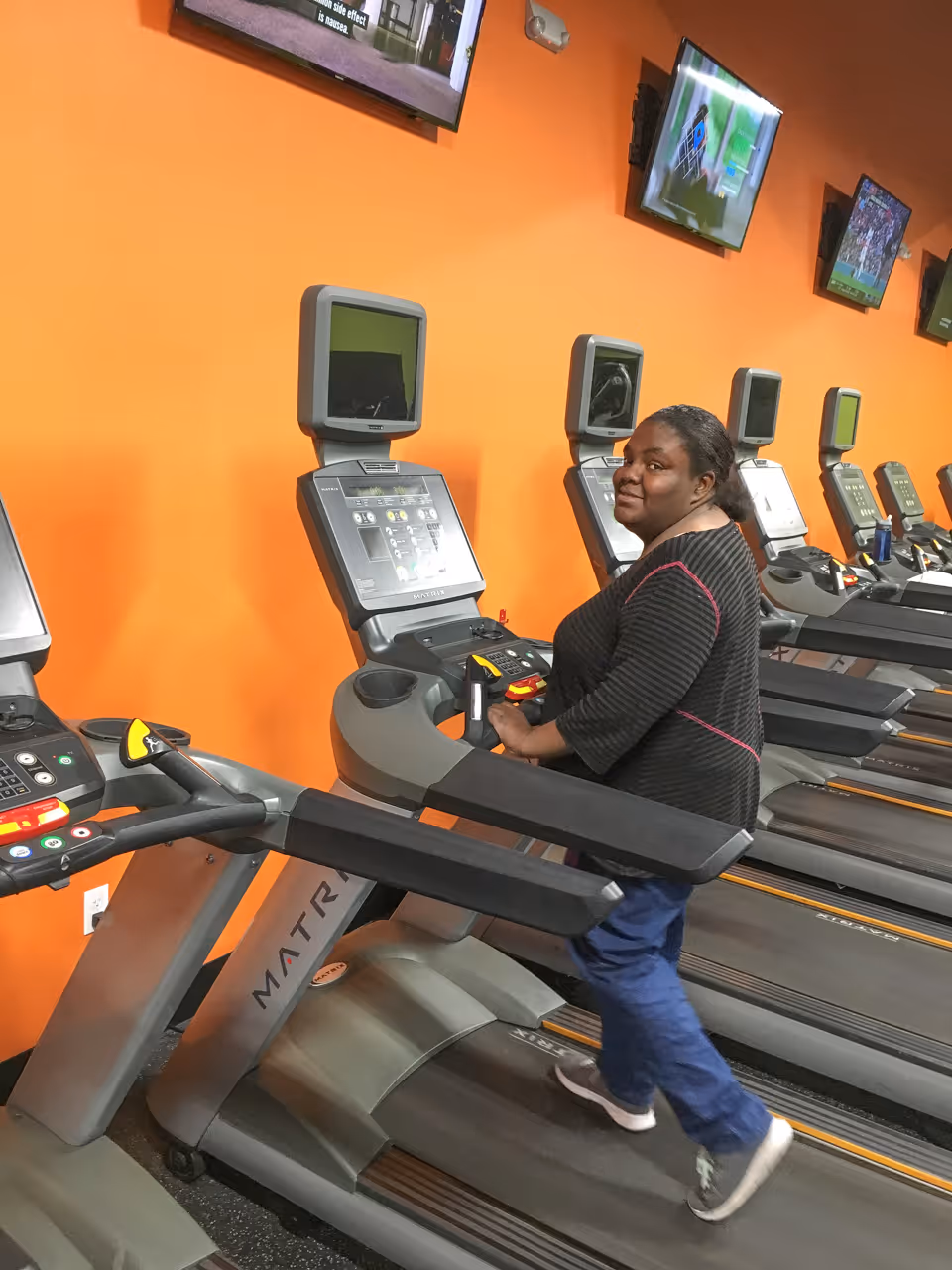 A woman walking on a treadmill in a gym with an orange wall and multiple treadmills lined up. Several televisions are mounted on the wall above the treadmills.