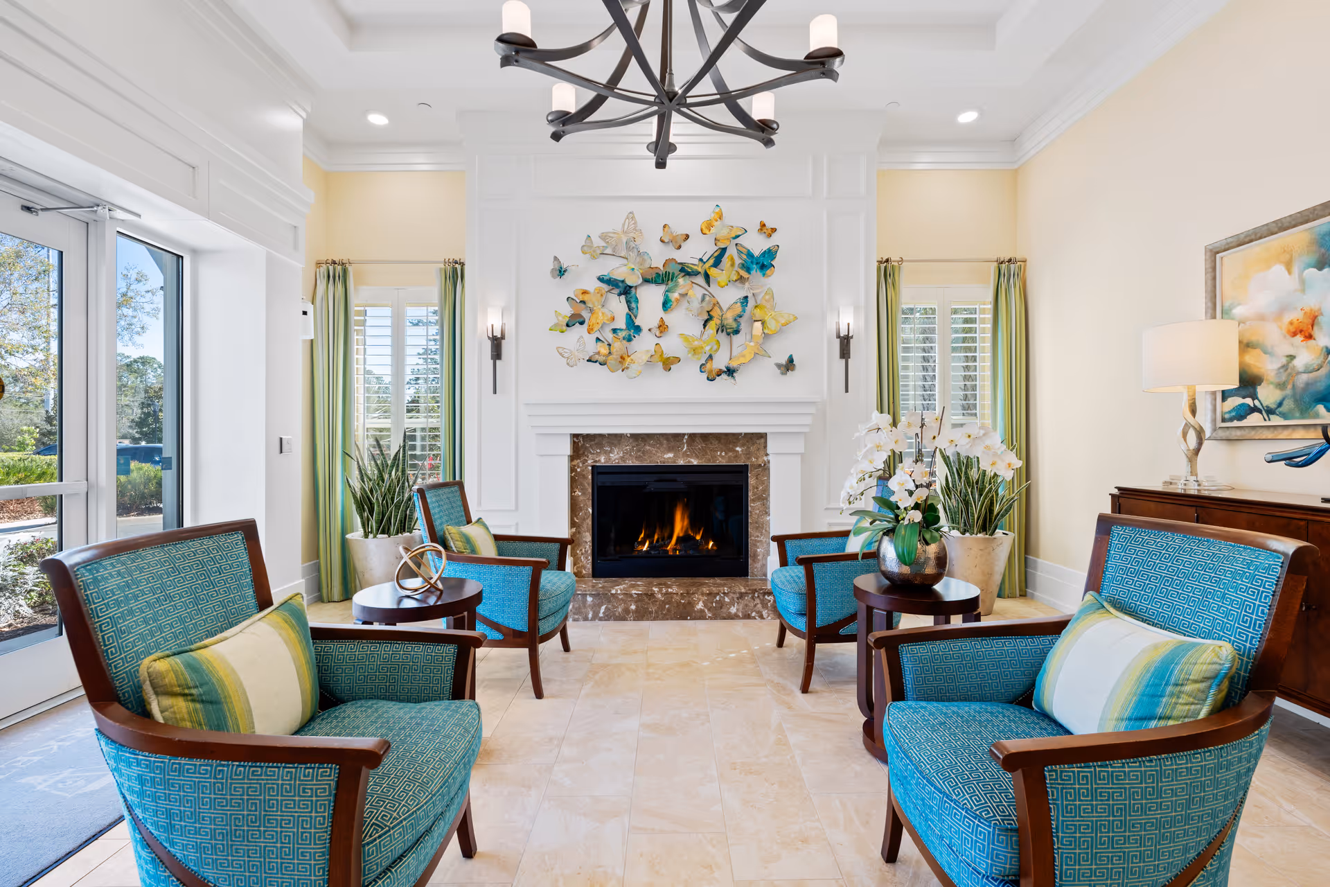 A bright and elegant living room with four turquoise armchairs arranged around a fireplace with a marble surround. Above the fireplace is a decorative wall art featuring colorful butterflies. The room has large windows with green curtains, potted plants, a chandelier, and a side table with a lamp and artwork on the wall.