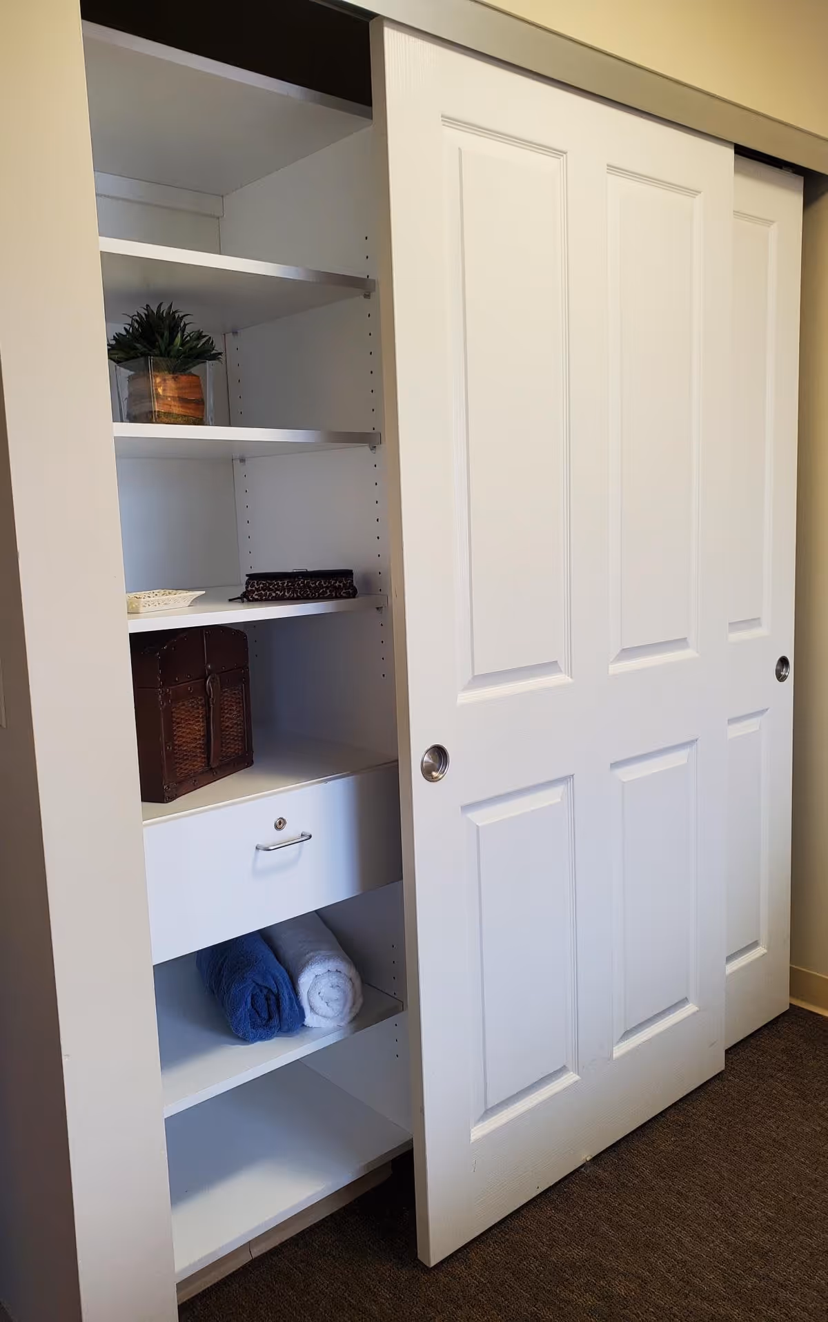 A white built-in closet with sliding doors, one door partially open revealing shelves inside. The shelves hold a small potted plant, a decorative box, a drawer, and two rolled towels, one blue and one white. The closet is set against a beige wall and carpeted floor.