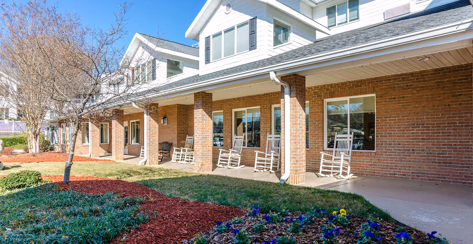 Exterior view of a senior living facility with a covered porch featuring several white rocking chairs. The building has brick walls and white siding on the upper level. There are landscaped garden beds with mulch, green shrubs, and some flowering plants in front of the porch. A leafless tree is visible on the left side under a clear blue sky.