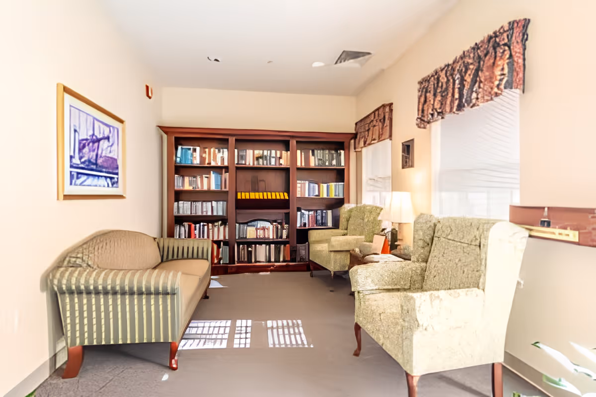 A cozy sitting area with a striped sofa on the left and two green upholstered armchairs on the right. A large wooden bookshelf filled with books is against the back wall. Two windows with patterned valances let in natural light, and a table lamp is placed between the armchairs.