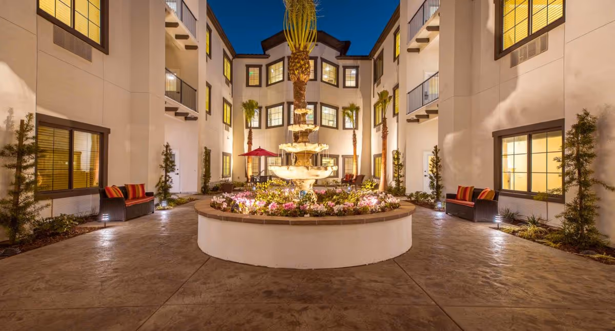 Nighttime courtyard with a central tiered fountain, flower beds, palm trees and seating surrounded by a multi-story senior living building.