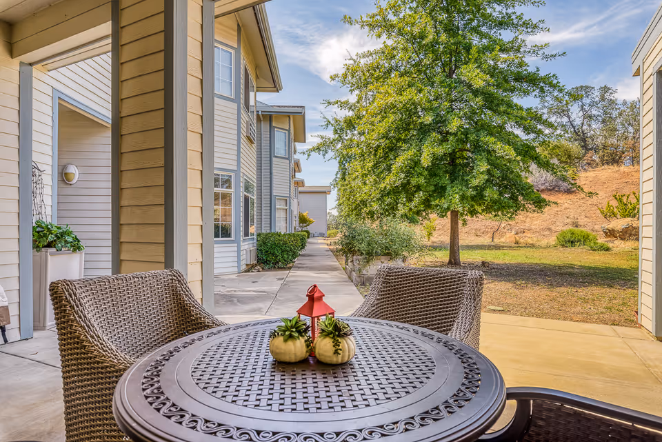 Shaded patio with a round metal table and wicker chairs overlooking a walkway, landscaping, and the exterior of a senior living building.