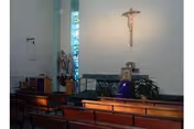 Interior view of a small chapel or prayer room with wooden pews, a crucifix mounted on the wall, a statue of a religious figure, and a lectern draped with a purple cloth. There is a stained glass window allowing light into the space.