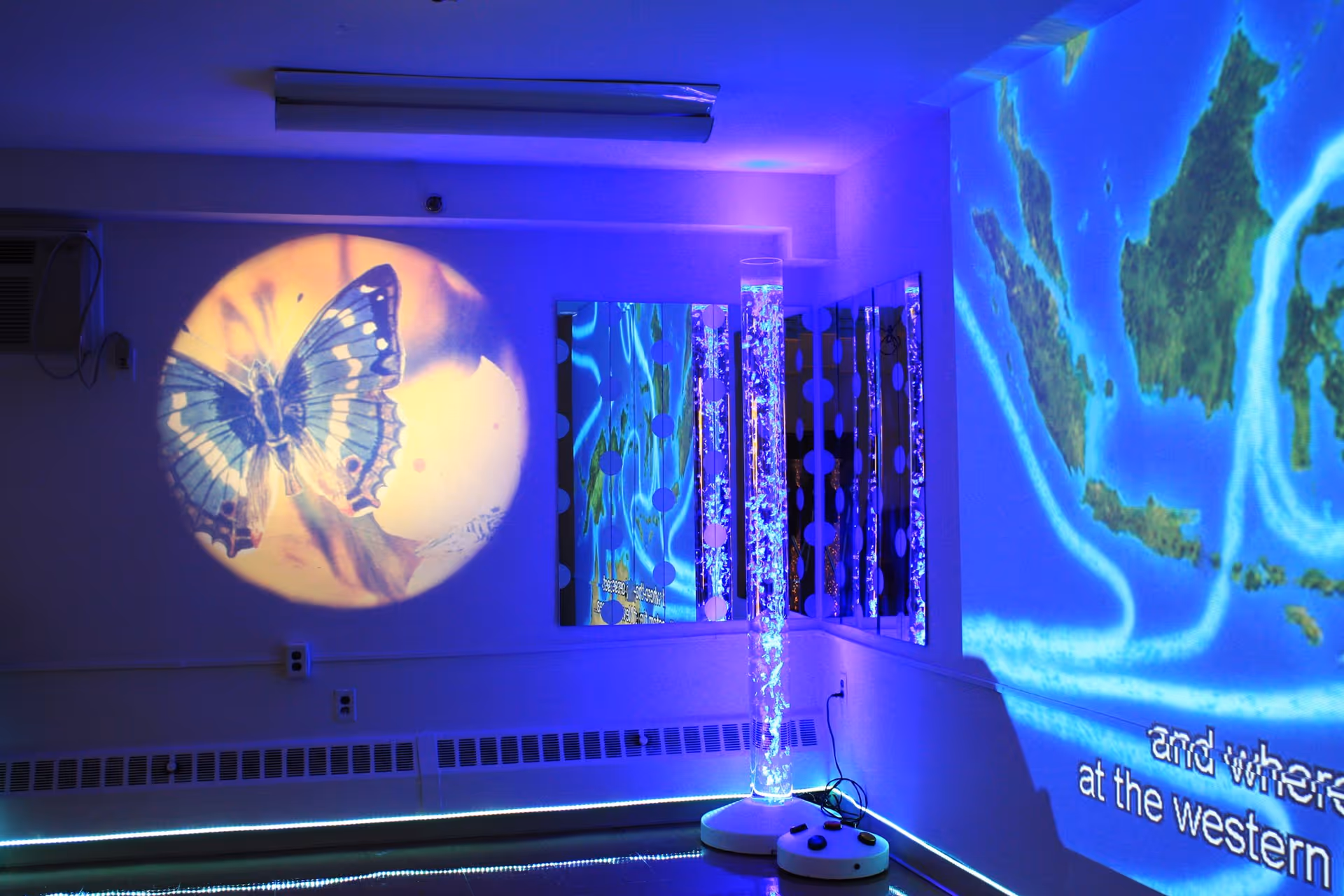 A sensory room with calming blue and purple lighting, featuring a projected image of a butterfly on one wall and a map on another. There is a tall bubble tube light in the corner with mirrors behind it, creating a soothing atmosphere.