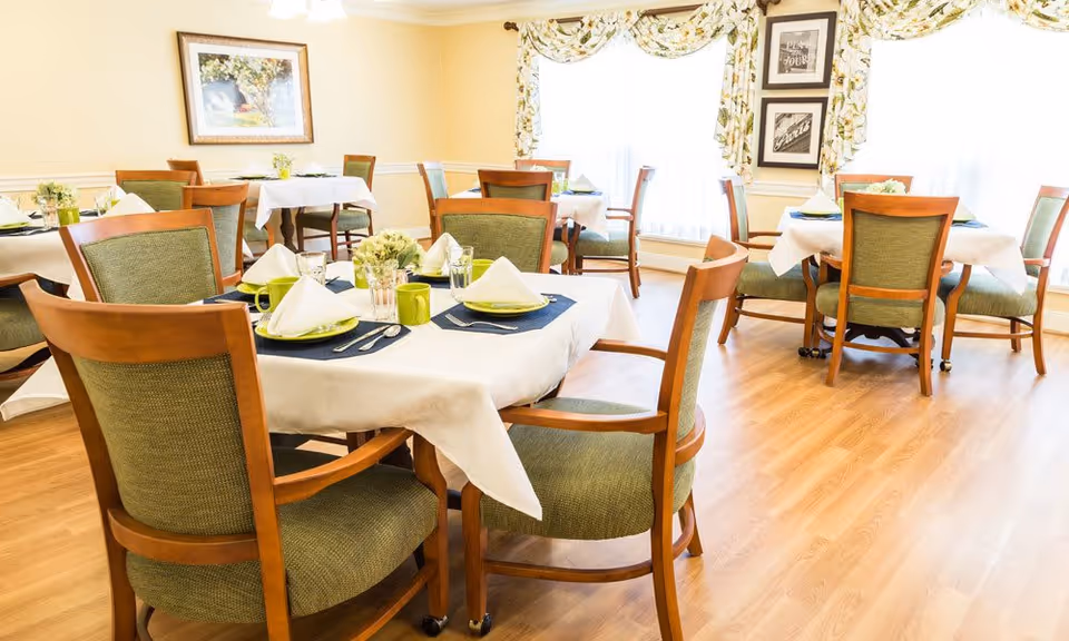 A bright dining room with several wooden tables covered with white tablecloths and set with green plates, cups, silverware, and napkins. The chairs have wooden frames and green upholstery. The room has light-colored walls, large windows with floral curtains, and framed artwork on the walls.