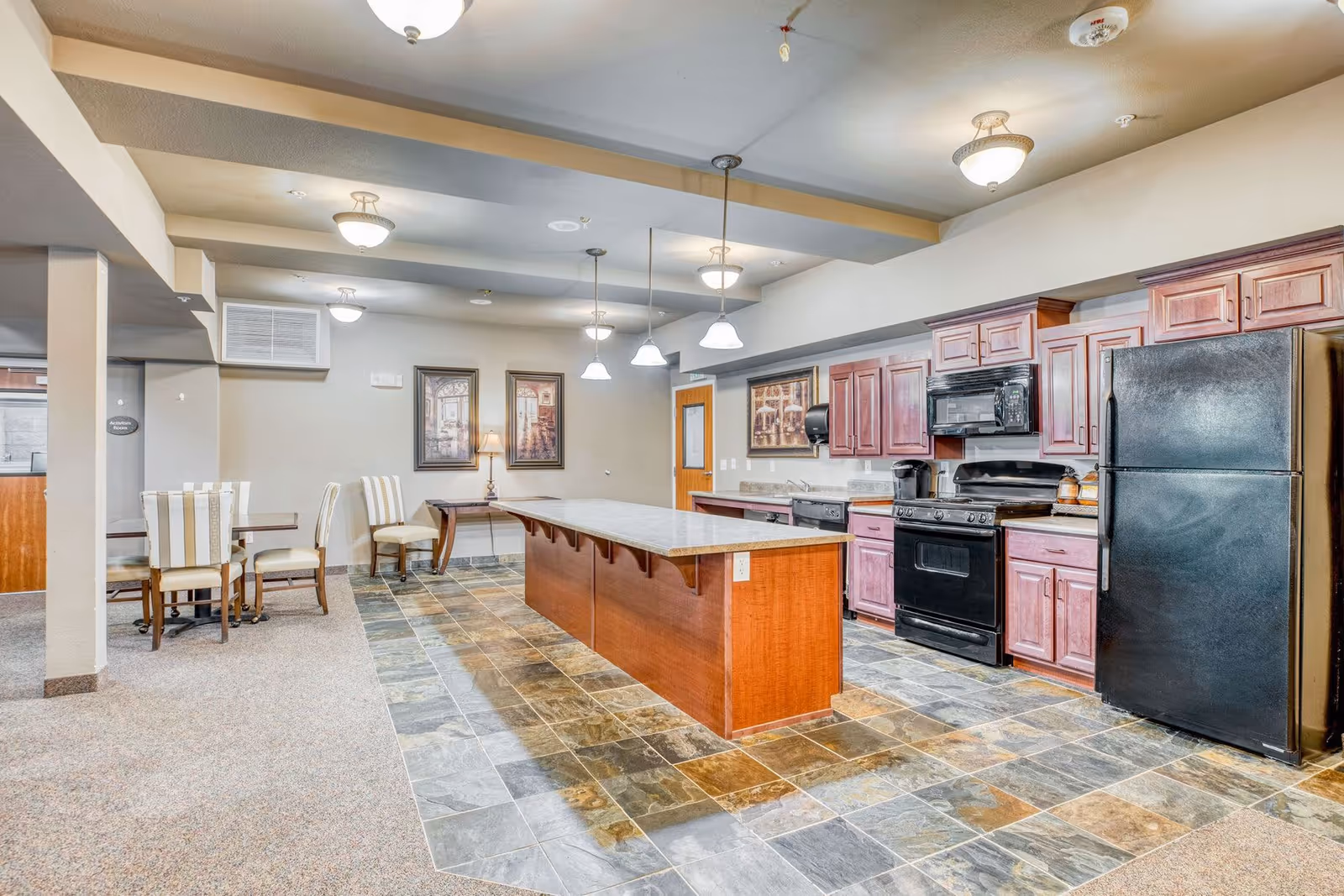 Large communal kitchen and dining area with a long center island, wood cabinetry, black appliances, and seating.