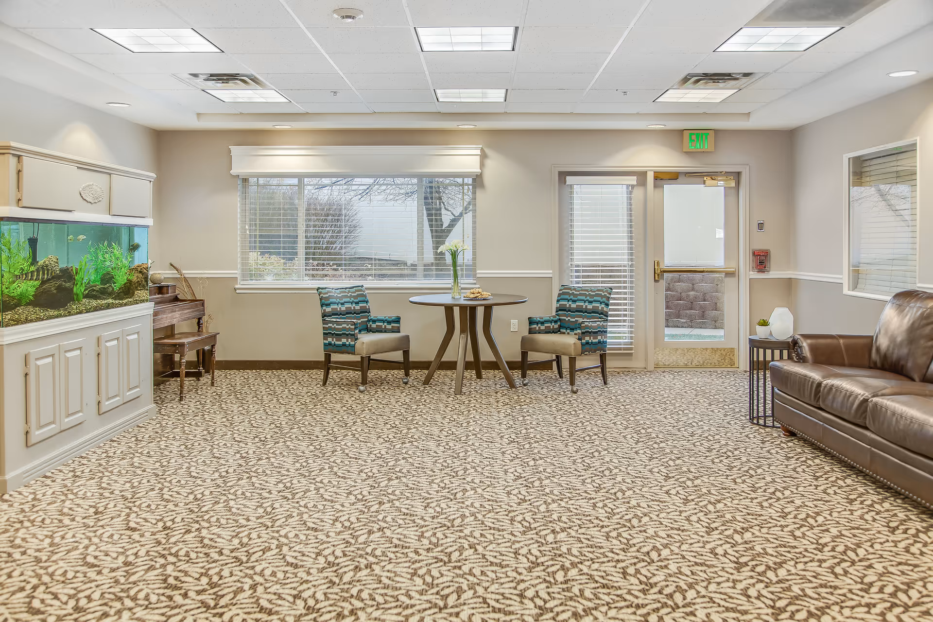 A well-lit common area with patterned carpet, two blue patterned chairs around a small round table with a vase of flowers and cookies, a large fish tank on the left, a piano behind the fish tank, a brown leather couch on the right, and a door and window with blinds at the back.