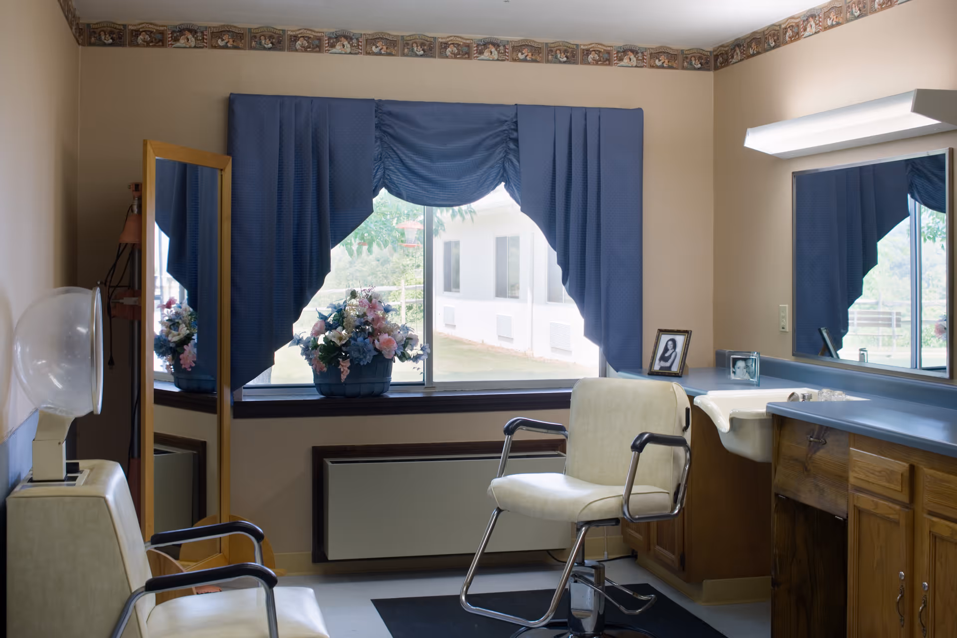 Small salon-style room with styling chairs, a sink and mirror, and a window with blue curtains and a floral arrangement.