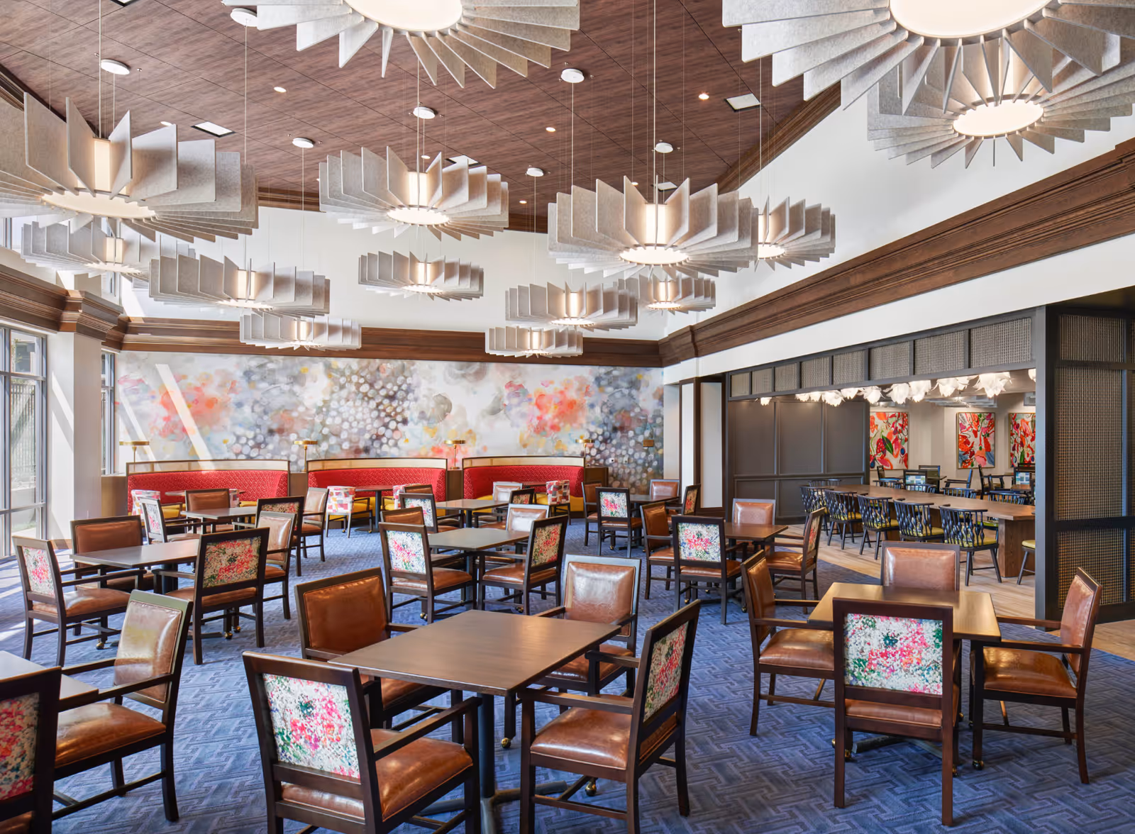 A spacious dining room with multiple wooden tables and chairs featuring floral upholstery. The room has a blue patterned carpet, red cushioned bench seating along the back wall, and large modern pendant lights hanging from a wooden ceiling. There is a colorful abstract mural on the far wall and an adjoining room visible through a large open doorway with additional seating and vibrant artwork.