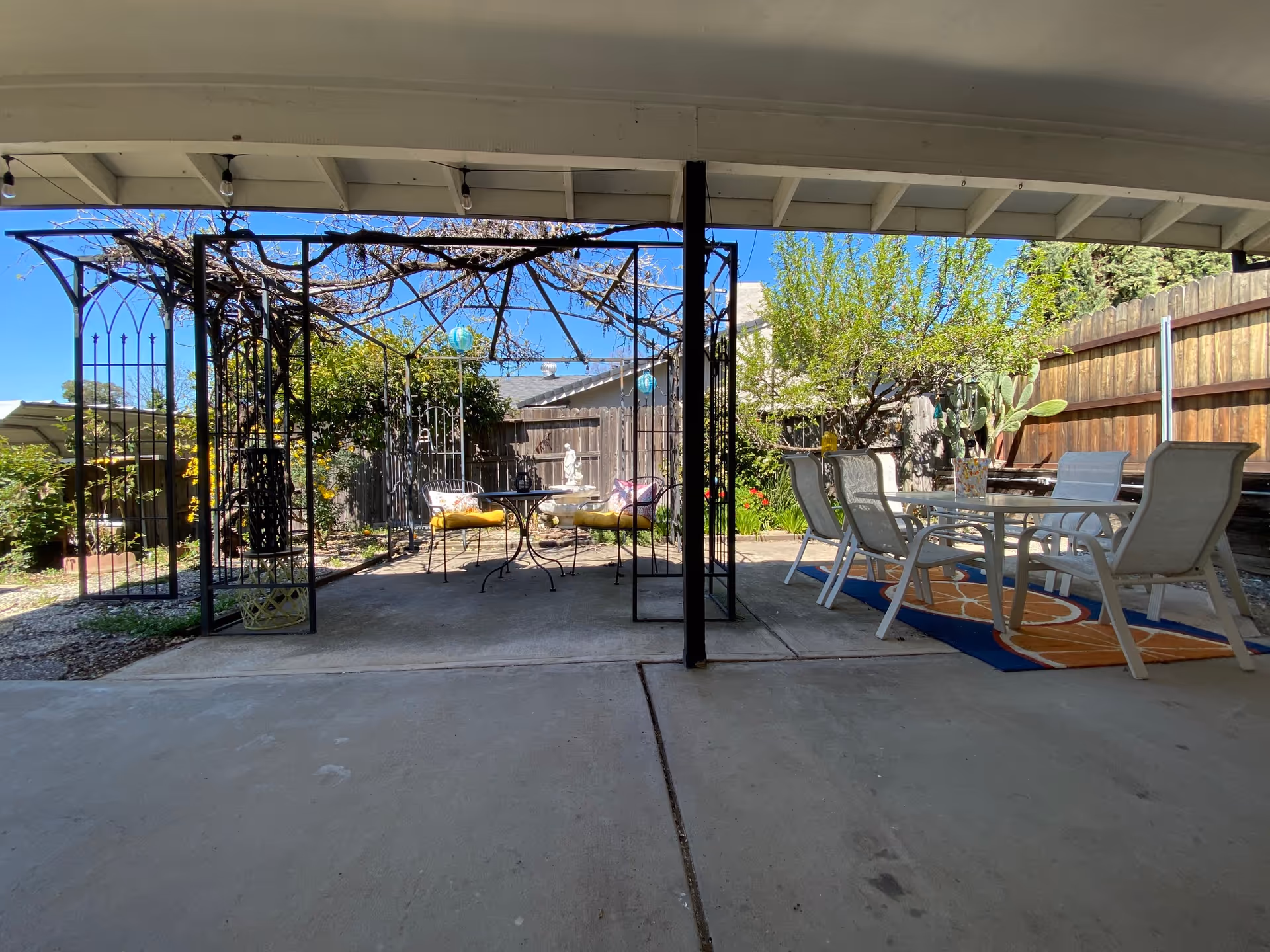 Outdoor covered patio area with a metal pergola structure on the left side featuring a small round table and two chairs with yellow cushions. On the right side, there is a glass-top table with six white chairs placed on a colorful outdoor rug. The area is surrounded by a wooden fence, trees, and plants under a clear blue sky.