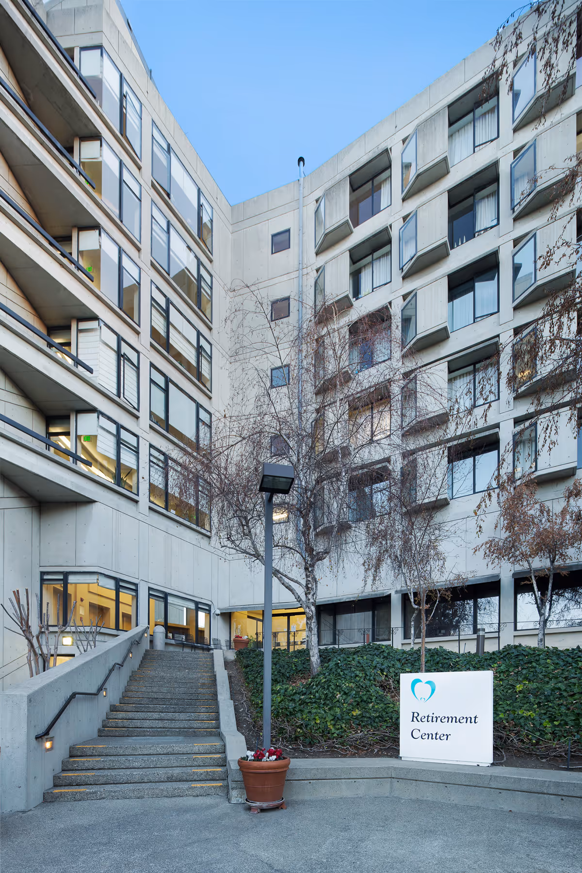 Exterior view of a multi-story retirement center building with large windows, a staircase leading to an entrance, a leafless tree, and a sign that reads 'Retirement Center' placed in front of some greenery.