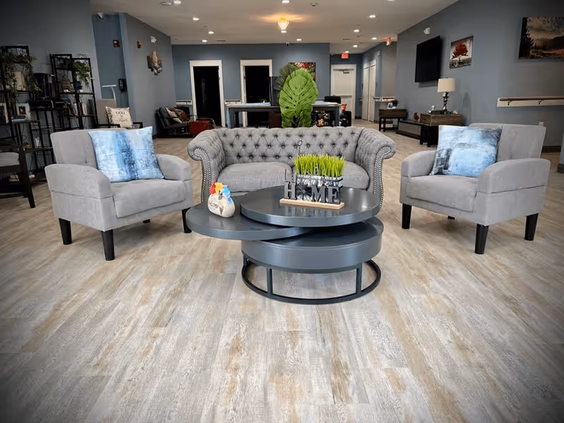A cozy living room area with a gray tufted sofa and two matching gray armchairs with blue pillows. In front of the seating is a modern round coffee table with decorative items including a small plant and a sign that says 'HOME'. The room has light wood flooring, blue-gray walls, and various pieces of furniture and artwork in the background.