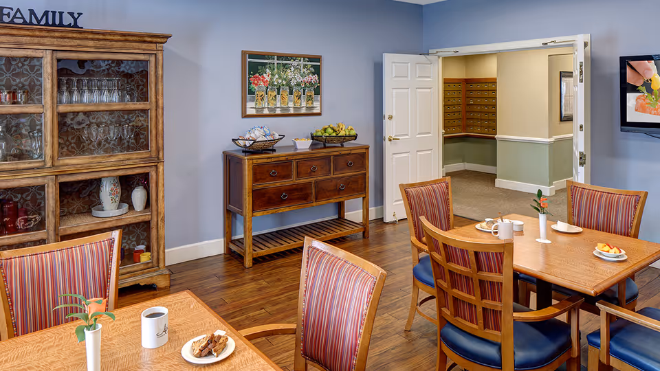 Dining room with wooden tables and striped chairs, a sideboard and glass-front cabinet against a blue wall, and an open door to a hallway.