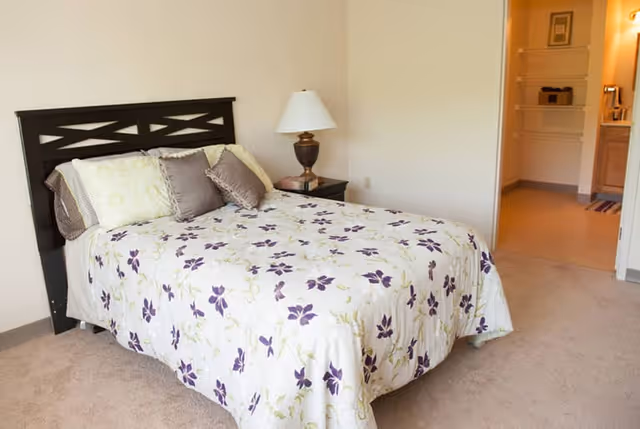 A bedroom with a bed covered in a white bedspread with purple floral patterns, two pillows, and a dark wooden headboard. There is a nightstand with a lamp next to the bed, and an open doorway leading to a closet and bathroom area.