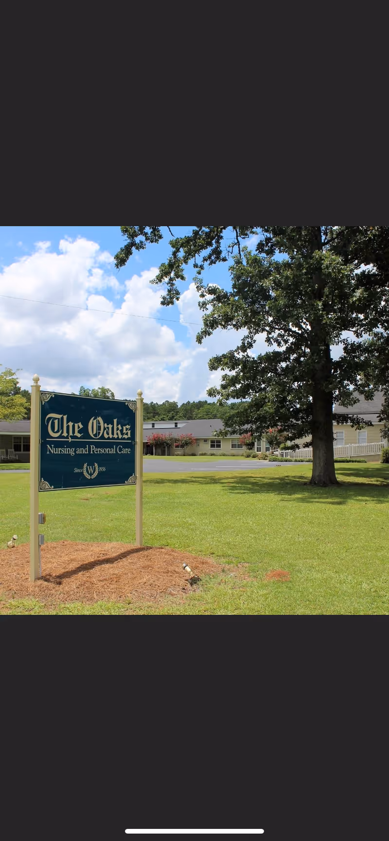 Outdoor view of The Oaks Nursing and Personal Care facility sign on a grassy lawn with a large tree and the building in the background under a partly cloudy sky.