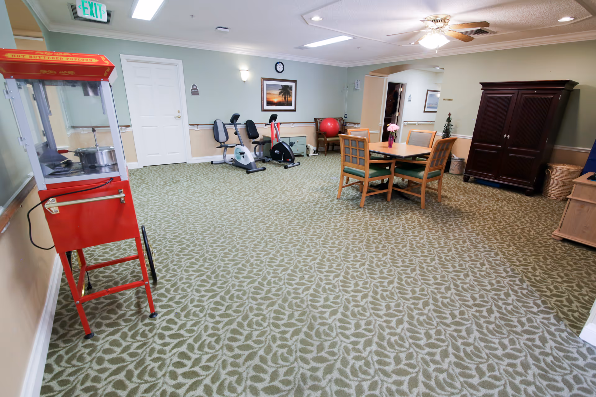A spacious room with patterned carpet featuring a red popcorn machine on the left, two exercise bikes against the far wall, a wooden table with four chairs and a vase of flowers in the center, and a dark wooden cabinet on the right. The walls are painted light green with beige wainscoting, and there is a ceiling fan with lights above. An exit sign is visible above a white door on the left wall.
