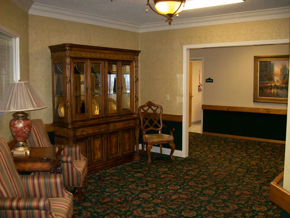 A sitting area in a senior living facility with two striped armchairs, a wooden side table with a decorative lamp, a wooden china cabinet displaying plates, and a wooden chair with an upholstered seat. The room has patterned carpet, beige walls with a dark green wainscoting, and a ceiling light fixture. A hallway with a framed painting and a green sign is visible in the background.