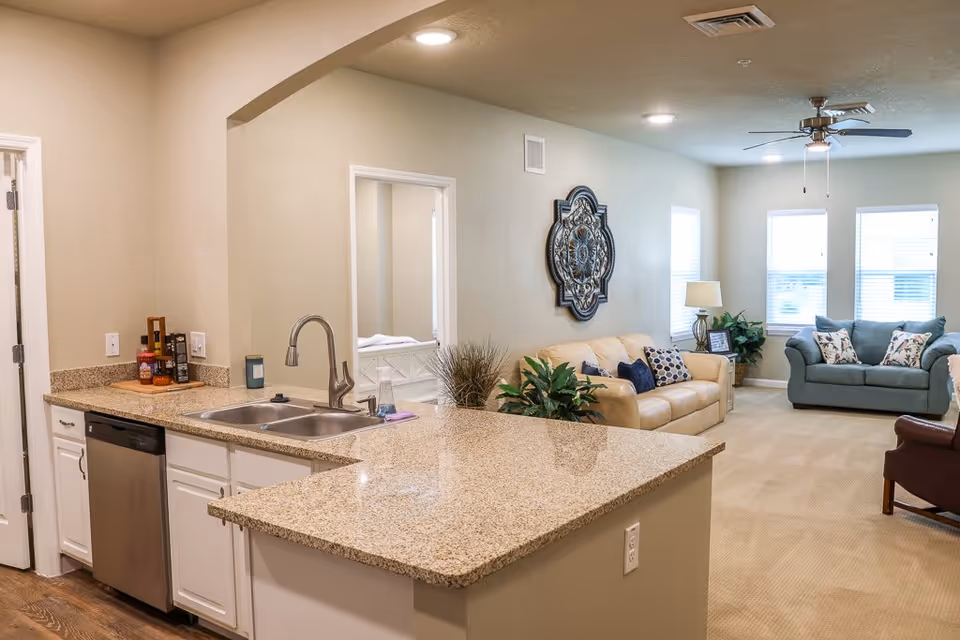 Interior view of a senior living facility showing a kitchen area with a granite countertop, double sink, dishwasher, and white cabinets. Beyond the kitchen is a living room with a beige sofa, a blue loveseat, decorative pillows, a ceiling fan, and three windows letting in natural light. The walls are painted a light beige color and there is a decorative wall piece above the sofa.