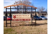 Outdoor view of a sign for Meridian Nursing & Rehabilitation Center located at 1555 N. Meridian with a phone number 942-8471. The sign is mounted on black metal posts with a wooden top frame. Behind the sign, there is a parking lot with several cars and a single-story building under a clear blue sky.