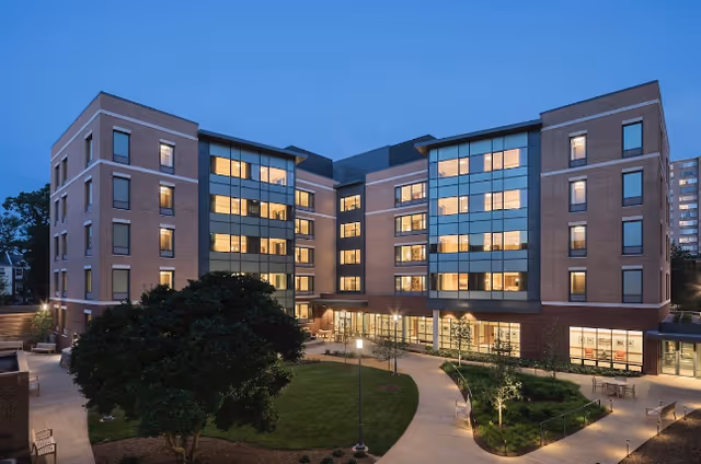 Exterior view of a modern multi-story senior living facility building at dusk with illuminated windows, landscaped garden, and paved walkways.