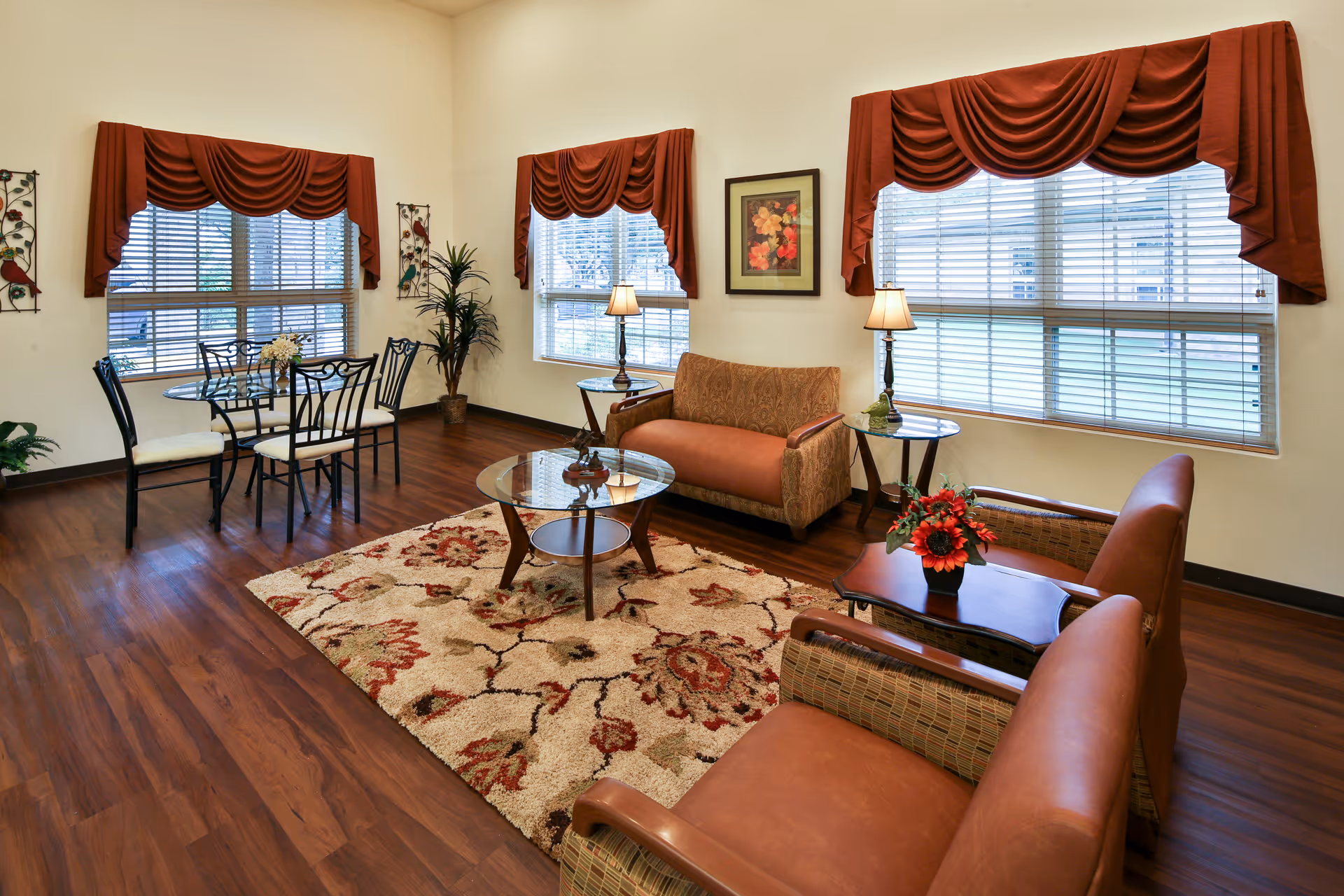 Well-lit common living area with sofas, armchairs, a glass coffee table, a dining set, and large windows with red valances.