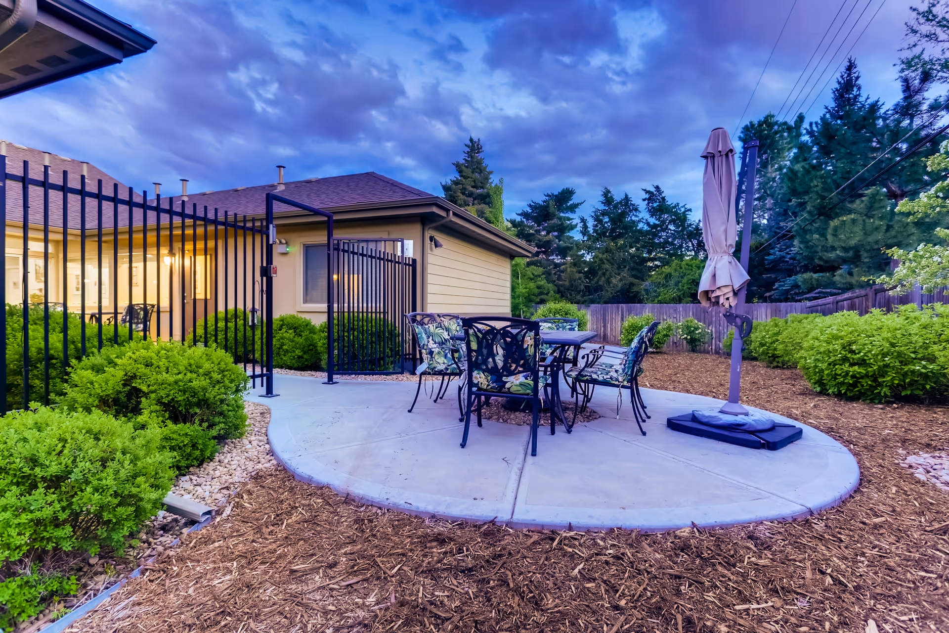 Outdoor patio area at Rocky Mountain Assisted Living and Memory Care with a round concrete seating area featuring a table and four chairs with floral cushions, surrounded by green bushes and trees under a cloudy evening sky.