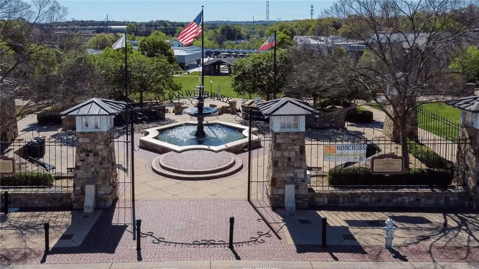 Gated outdoor plaza with a central fountain, stone pillars, flags, and surrounding trees.