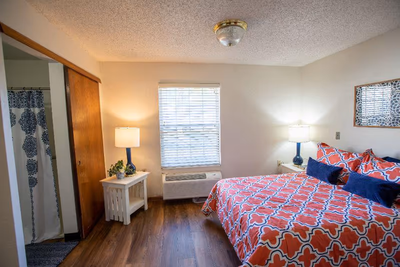 A cozy bedroom with a bed covered in a red and white patterned bedspread with blue accent pillows. There are two bedside tables with lamps on each side of the bed. A window with blinds is centered on the wall opposite the bed. To the left, there is an open doorway leading to a bathroom with a shower curtain featuring a blue and white pattern. The floor is wooden, and the walls are painted white.