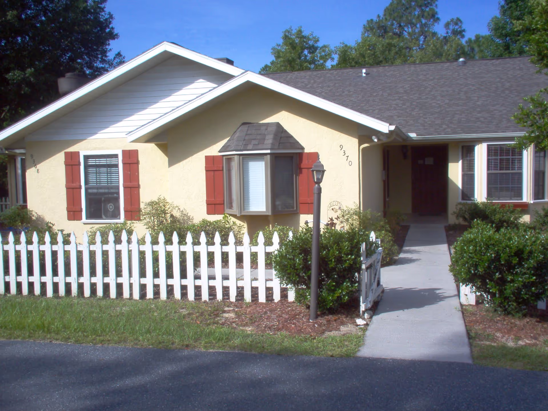 Exterior view of a single-story yellow house with red shutters and a white picket fence. There is a concrete walkway leading to a covered entrance, surrounded by green bushes and trees under a clear blue sky.