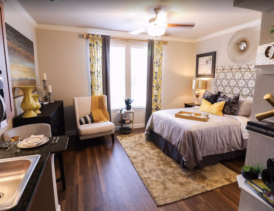 A cozy bedroom with a large bed adorned with gray and yellow pillows and a gray bedspread. There is a beige armchair with a yellow throw blanket near a window with yellow and gray patterned curtains. A small side table with a plant and books is placed by the window. The room features wooden flooring, a patterned rug, a lamp on a nightstand, and decorative wall art. A kitchen counter with a sink and place settings is visible in the foreground.